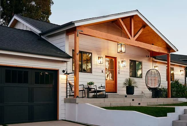 White craftsman home at dusk with a wood-beam porch, orange front door, and warm exterior lighting.