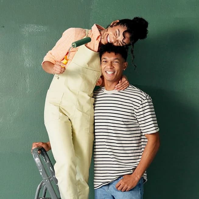 A smiling couple painting a dark green wall, one standing on a step ladder holding a paint roller.