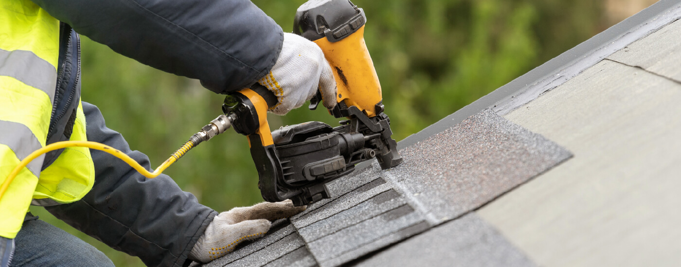 A closeup photo shows someone kneeling on top of a roof while using a tool to lay down new shingles.