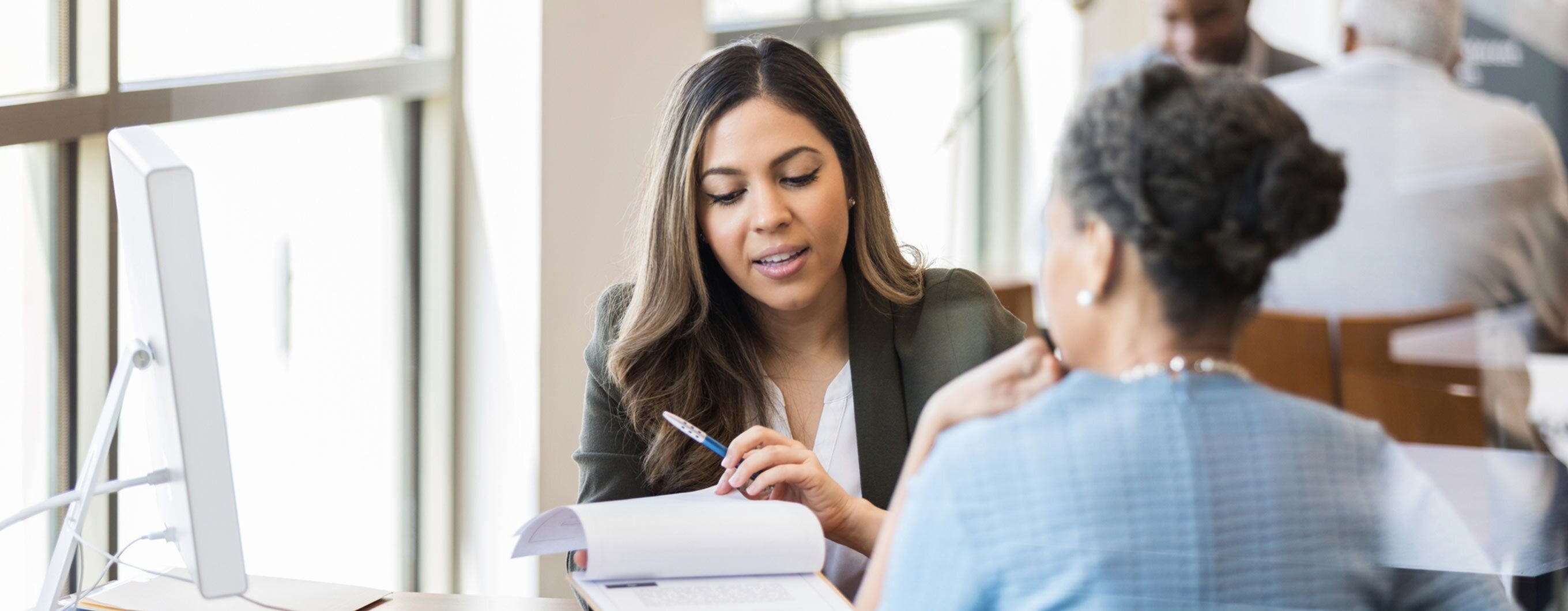 Two women sit at a desk, one facing the camera and the other facing away. The woman facing the camera is holding a clipboard.
