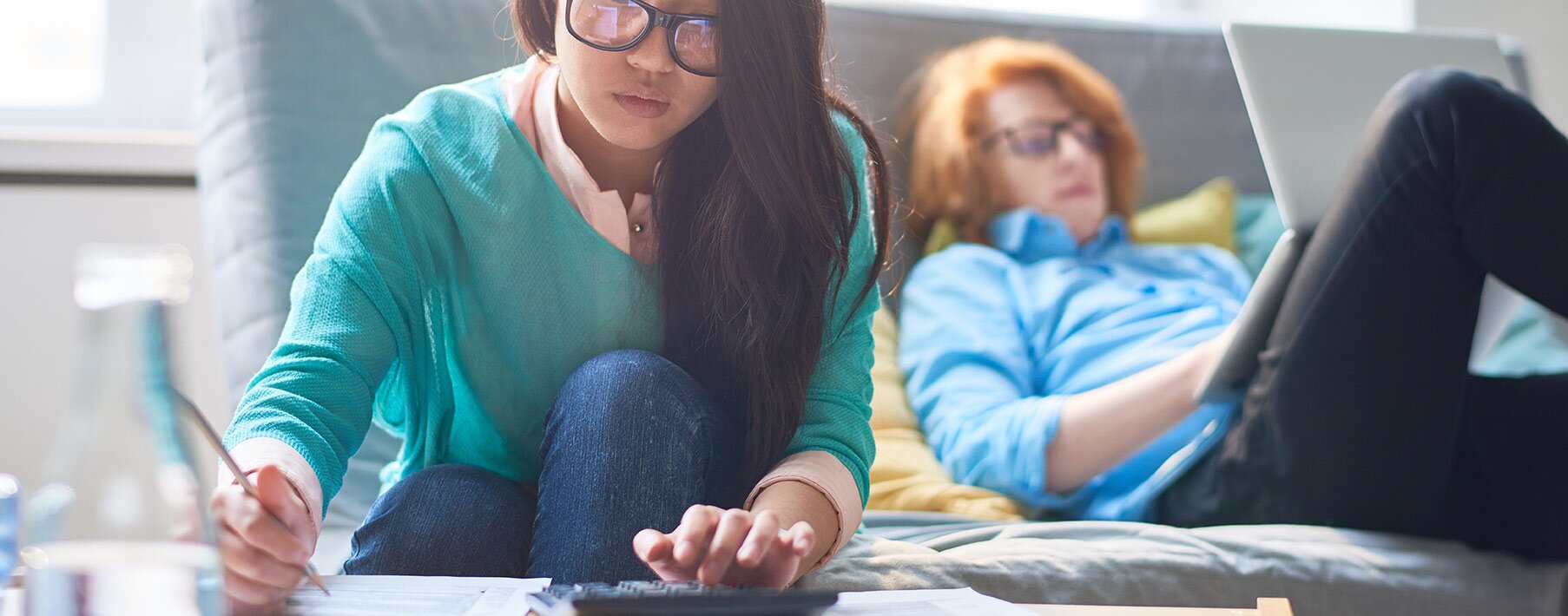 Two people on a couch, one leaning forward and using a calculator and the other leaning back and using a laptop