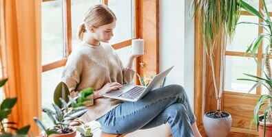 A woman sitting against a window surrounded by plants is holding a mug and working on a laptop