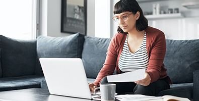 A young woman sitting on a couch looking at her laptop and a piece of paper