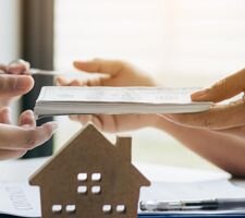 Image of hands passing money from one to another with a wooden cut out of a home in front of them
