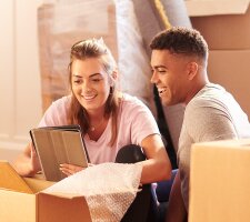 A young couple sits on the floor looking at a tablet surrounded by moving boxes