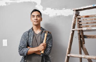 A man wears safety gear while working on a piece of drywall with a large crack in it, alluding to the topic of how to deal with cracks in the walls of your home.