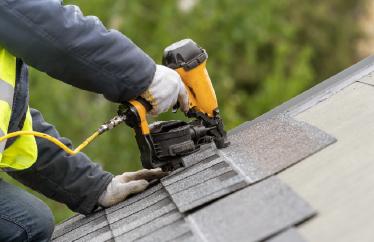 A closeup photo shows someone kneeling on top of a roof while using a tool to lay down new shingles.